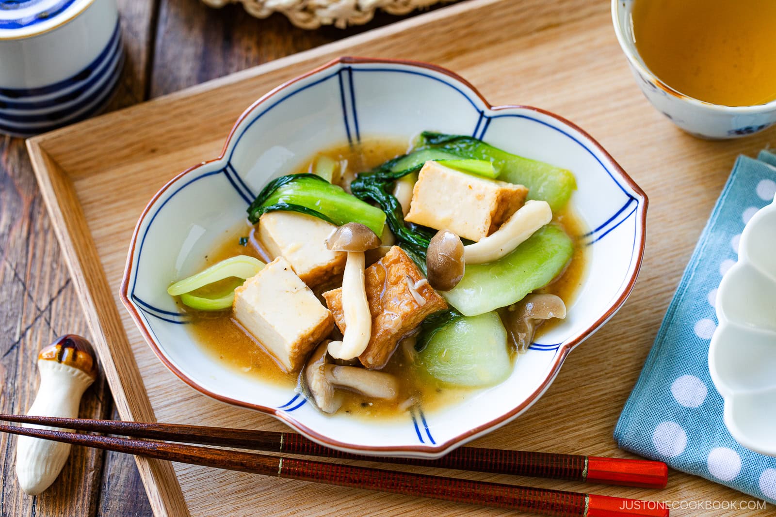 A bowl of bok choy and tofu stir-fry with mushrooms and savory sauce is served on a wooden tray with chopsticks and a cup of tea, beautifully presented in a white and blue flower-shaped bowl.
