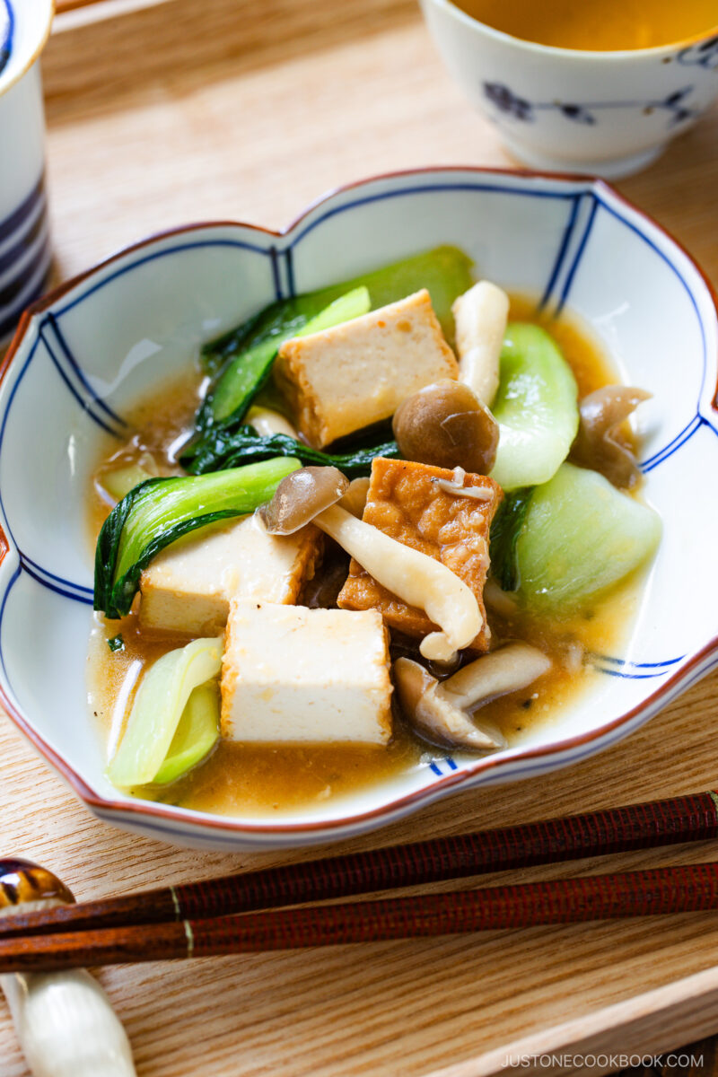 A bowl of bok choy and tofu stir-fry with assorted mushrooms in a light brown sauce, served in a decorative dish on a wooden tray, accompanied by chopsticks and a cup of tea.