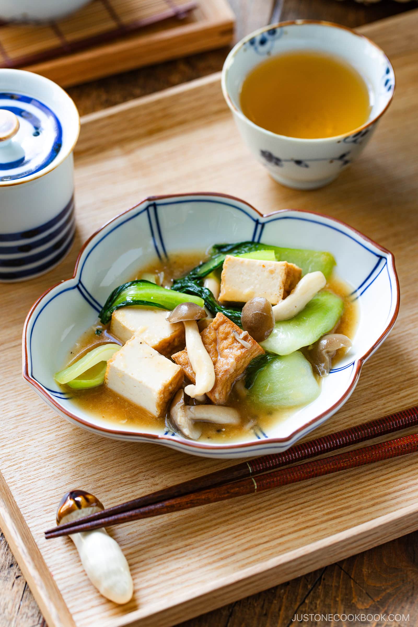 A bowl of bok choy and tofu stir-fry, paired with mushrooms in a light broth, is served on a wooden tray with chopsticks. A cup of tea and a small lidded bowl complete this elegant presentation.