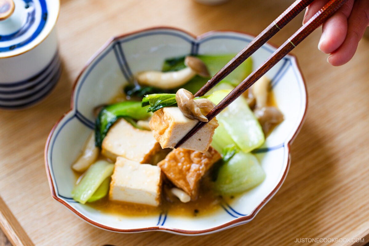 A hand with chopsticks picks up tofu from a savory bowl of bok choy and tofu stir-fry, featuring braised vegetables and mushrooms. The dish rests on a wooden surface beside a lidded cup.