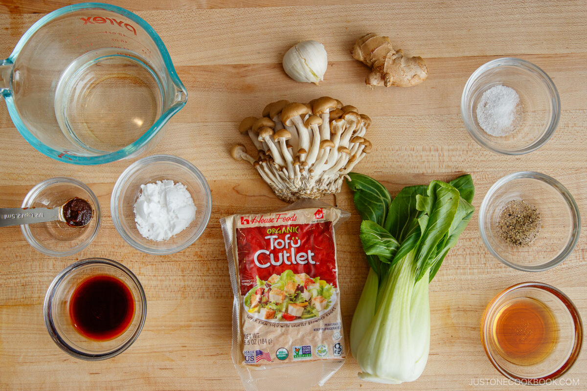 Ingredients for a flavorful bok choy and tofu stir-fry on a wooden surface: water in a measuring cup, garlic, ginger, shimeji mushrooms, baby bok choy, tofu cutlet, soy sauce, cornstarch, miso paste, salt, pepper, and sesame oil in small bowls.