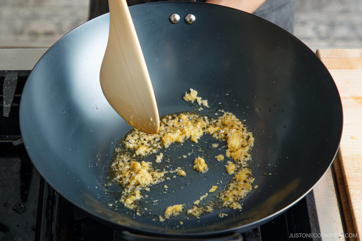 Minced garlic and ginger being sautéed in oil in a black wok on a stovetop, stirred with a wooden spatula as the base for a flavorful bok choy and tofu stir-fry.