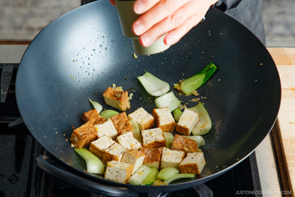 A hand sprinkles pepper from a grinder onto a bok choy and tofu stir-fry sizzling in a black wok on the stovetop.