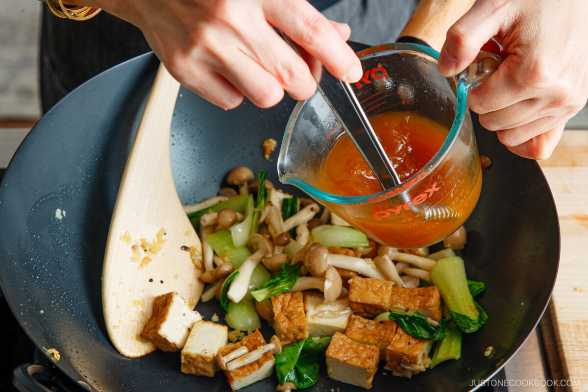 A person pours sauce from a measuring cup into a wok filled with bok choy and tofu stir-fry, mushrooms, and other vegetables while stirring with a spatula.