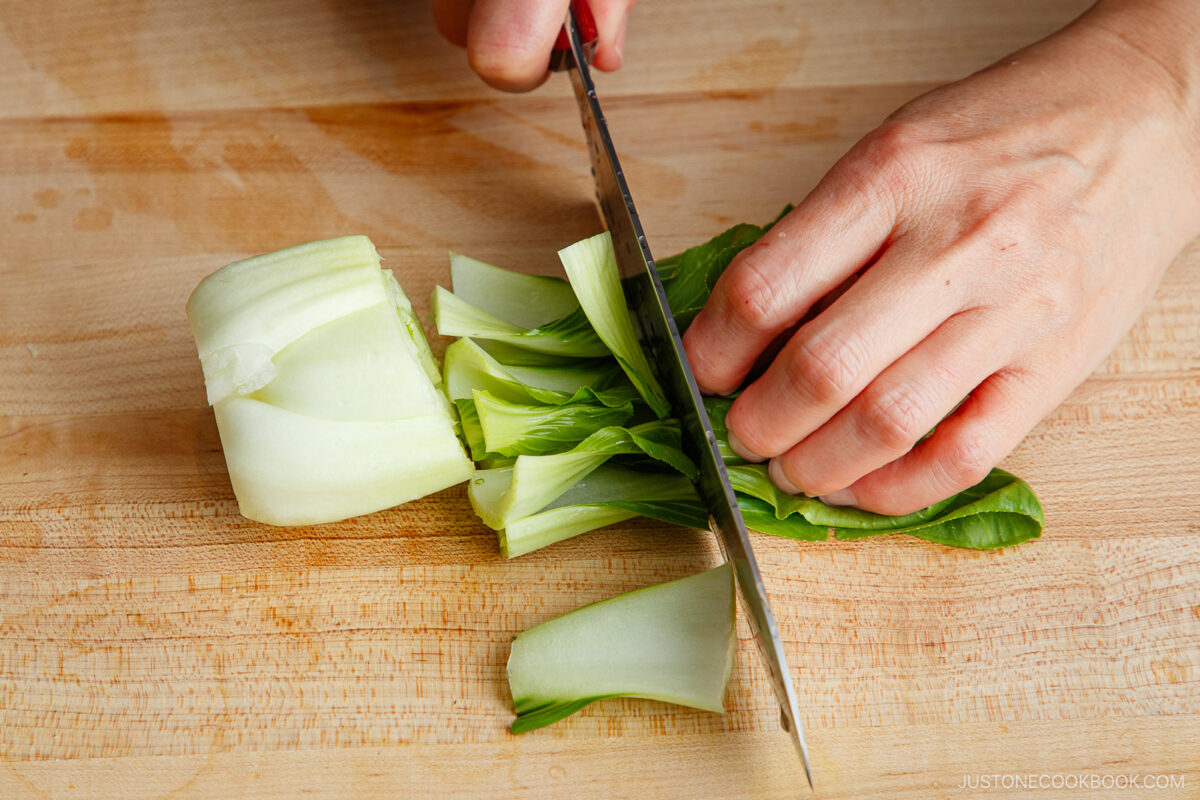 A person slicing a head of bok choy on a wooden cutting board with a large kitchen knife, prepping fresh ingredients for a flavorful bok choy and tofu stir-fry.