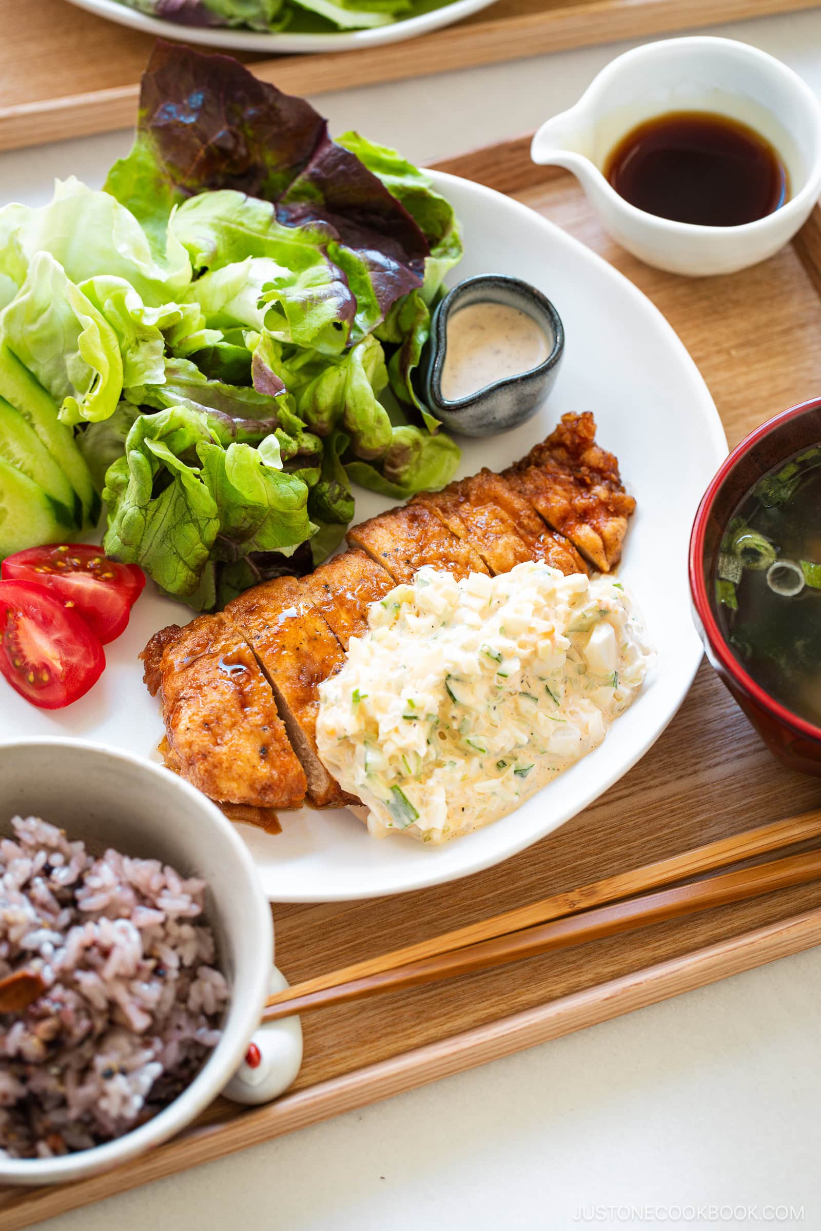 A plate of crispy fried chicken nanban topped with creamy sauce, fresh lettuce, cucumber, and tomato, with a small dish of dressing. Served with a bowl of mixed rice, soup, and dipping sauce on a wooden tray.