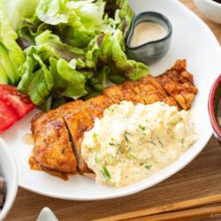 A plate of crispy chicken nanban topped with creamy egg salad, served alongside fresh lettuce, cucumber, tomato, a small dish of sauce, a bowl of miso soup, and multigrain rice.