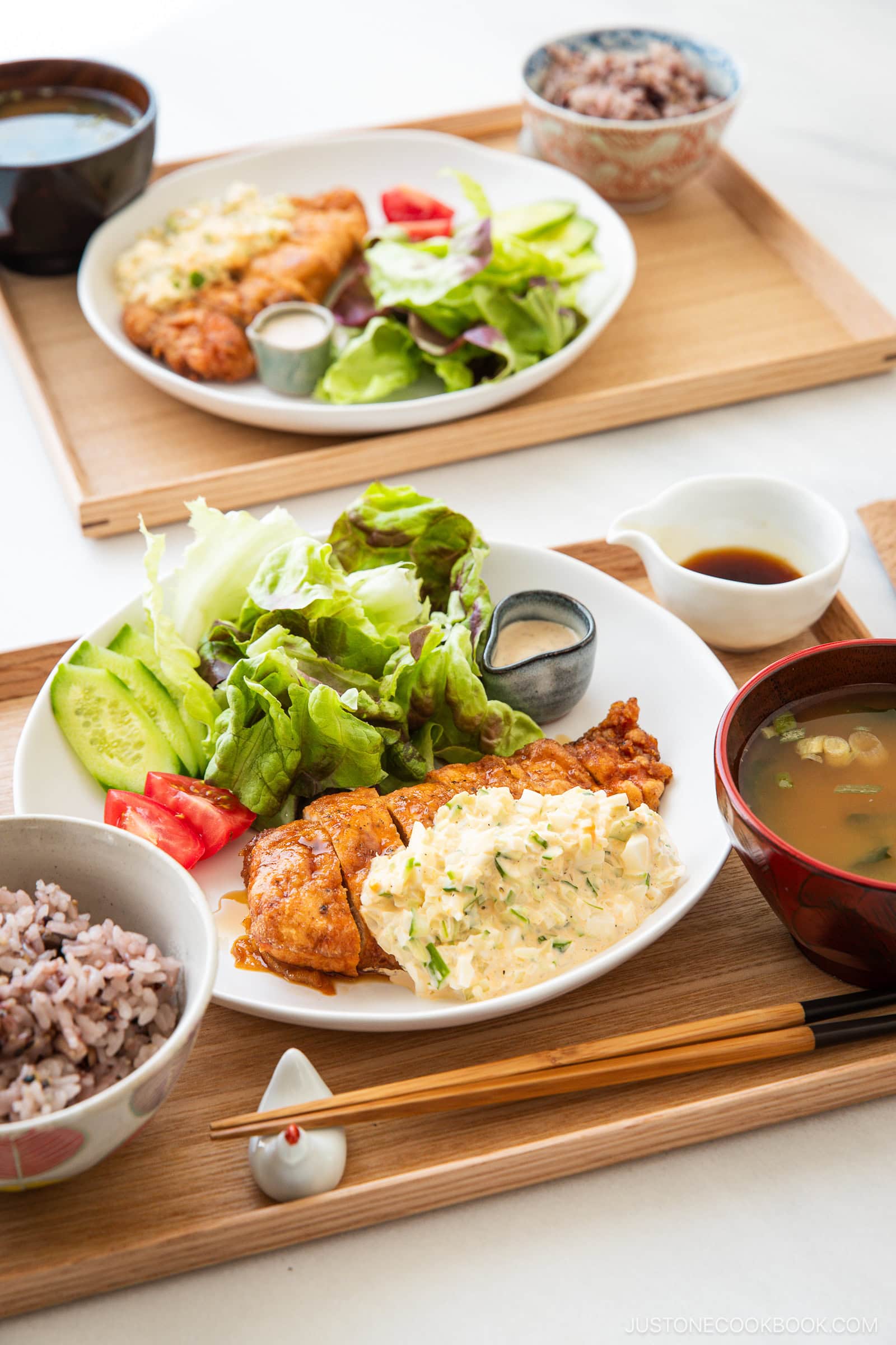 Two trays with plates of crispy chicken nanban topped with tartar sauce, fresh salad, tomato, cucumber, small bowls of miso soup, multigrain rice, and dressing, all arranged neatly with chopsticks on the side.
