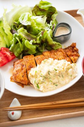 A plate with sliced chicken nanban topped with creamy egg salad, fresh lettuce, cucumber, and tomato slices. A small dish of dressing and wooden chopsticks are placed on the tray next to the plate.