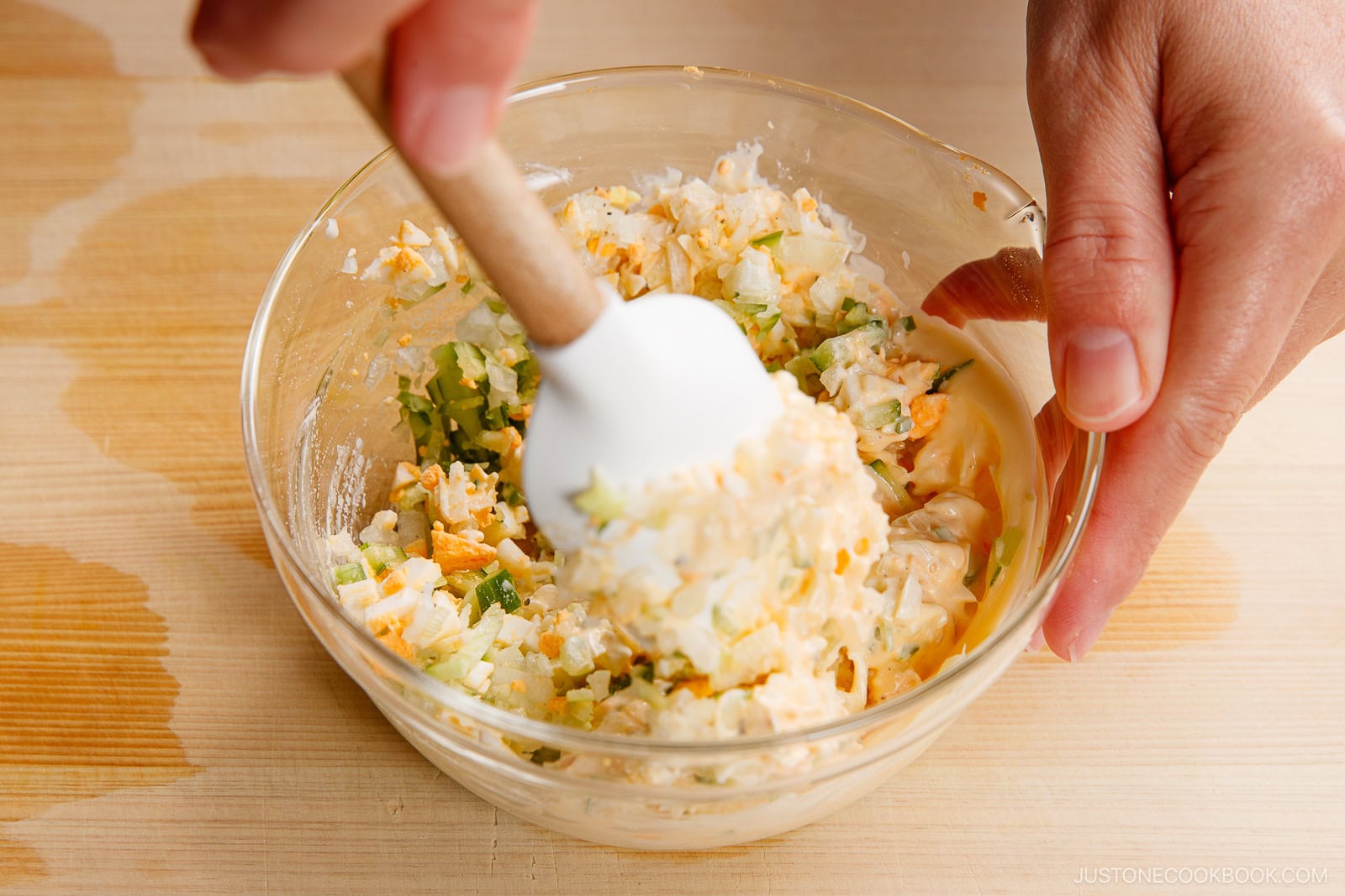 A hand uses a spatula to mix chopped eggs, celery, and mayonnaise in a glass bowl on a wooden surface—creating a base similar to chicken nanban sauce—with some spilled liquid around the bowl.