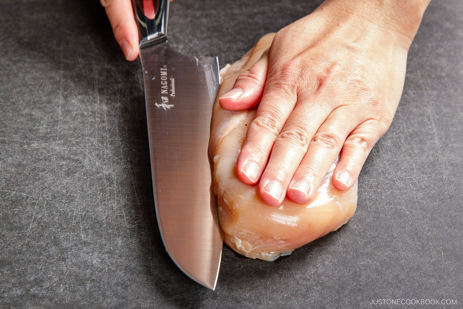 A person holds a raw chicken breast with one hand while slicing it horizontally with a large kitchen knife on a dark cutting board, preparing the meat for chicken nanban.