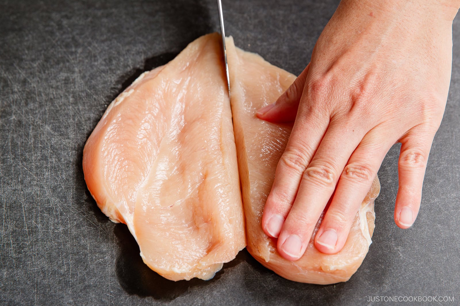 A hand holding a raw chicken breast on a dark cutting board while slicing it in half lengthwise with a knife, preparing the meat for chicken nanban.