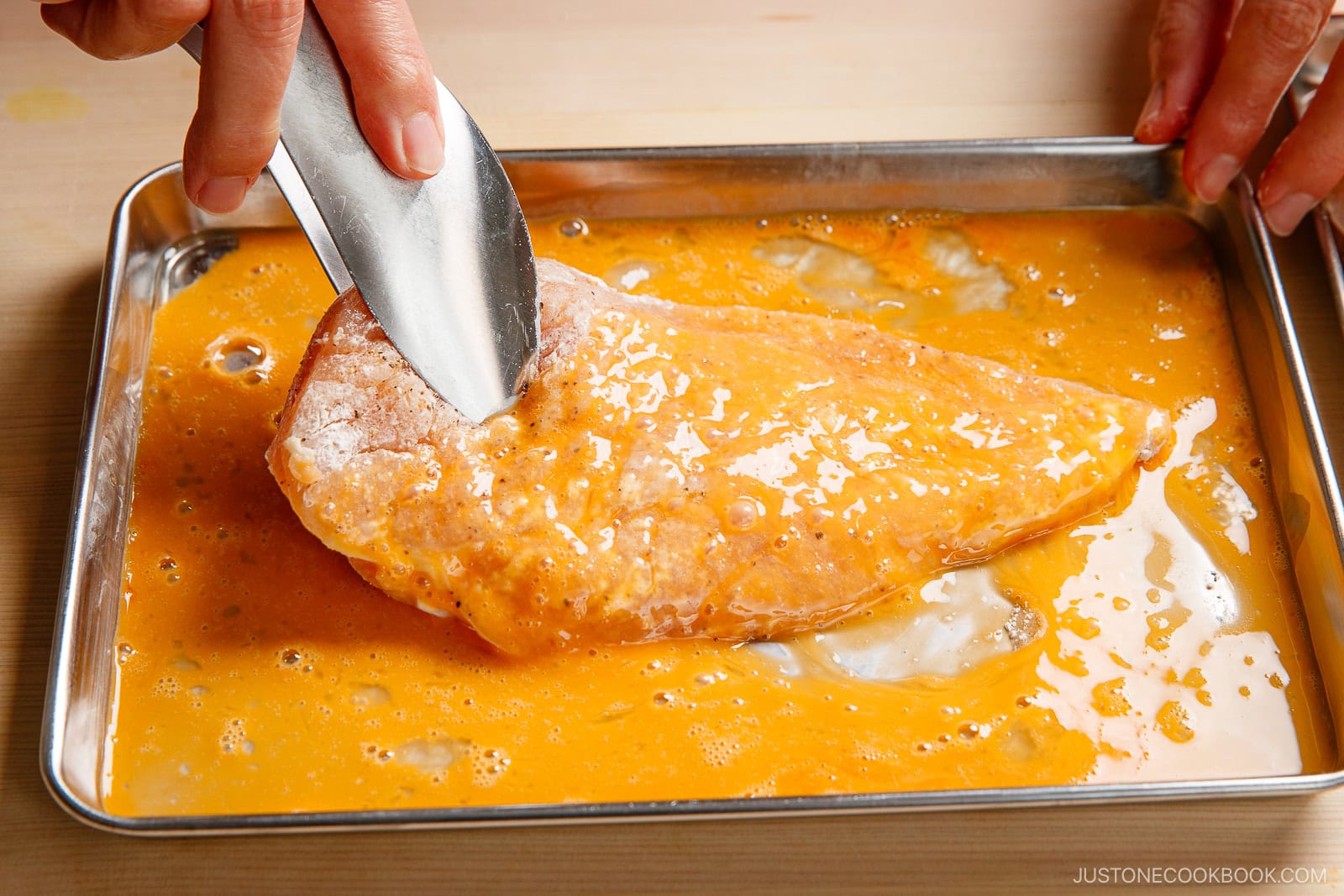 A hand uses tongs to dip a flour-coated fish fillet into a tray of beaten eggs, preparing it for breading or frying—similar to the method used in making chicken nanban.