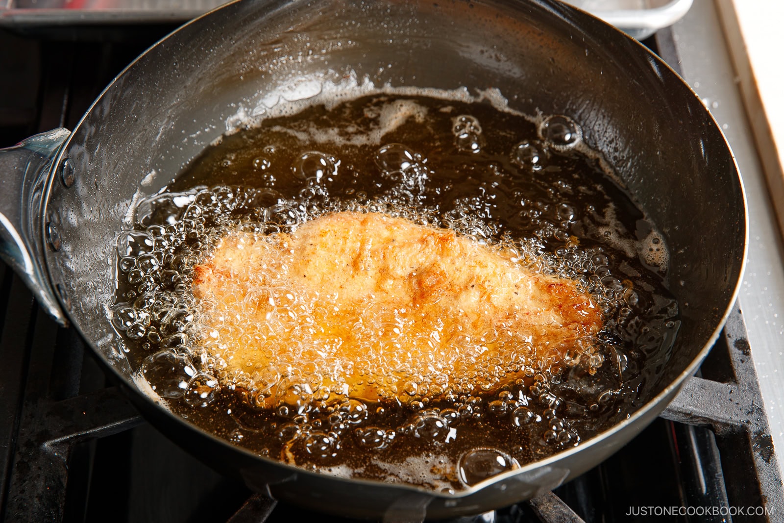 A breaded fish fillet, reminiscent of chicken nanban, is being deep-fried in hot oil in a black frying pan on a stovetop, with bubbles forming around the fillet.