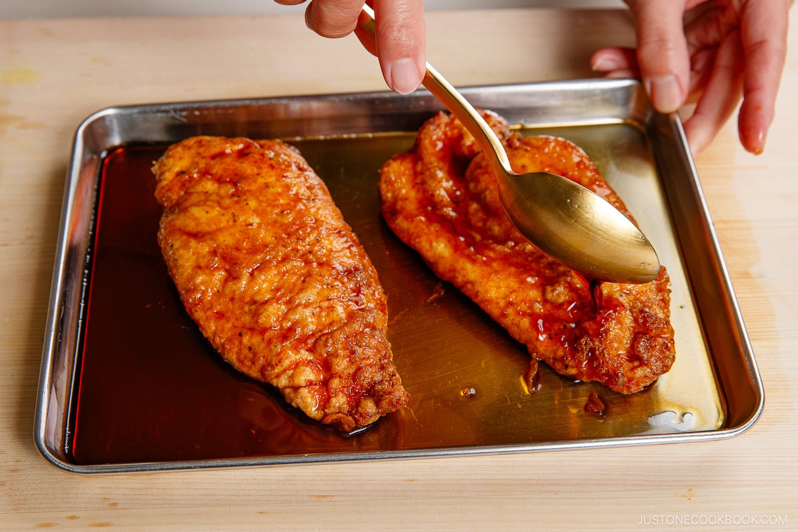 A hand uses a gold spoon to drizzle tangy sauce over two cooked, seasoned chicken nanban breasts on a metal tray.