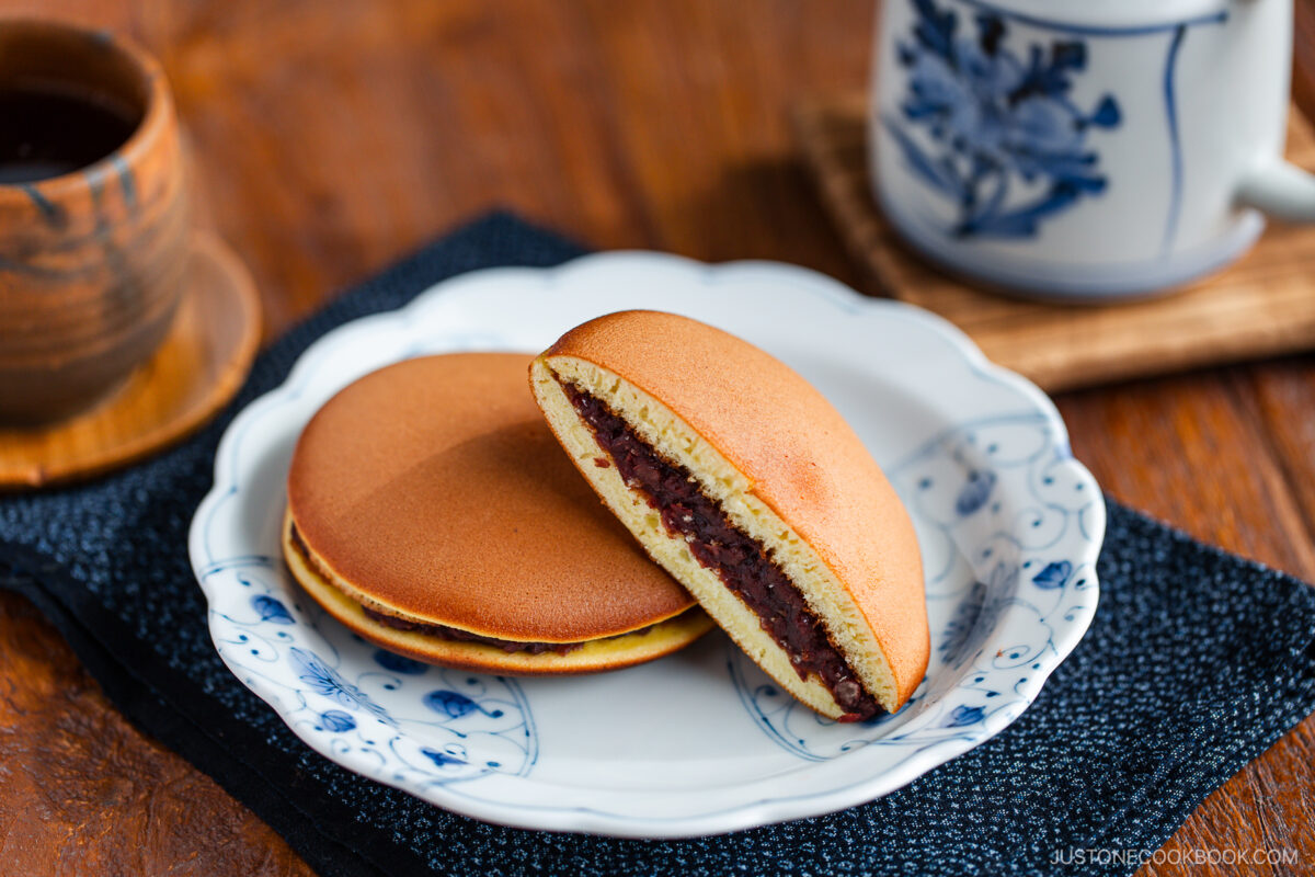 Two dorayaki (Japanese red bean pancakes) sit on a white and blue plate; one is whole, and the other reveals its sweet red bean filling. In the background, a cup and teapot rest on a wooden table.