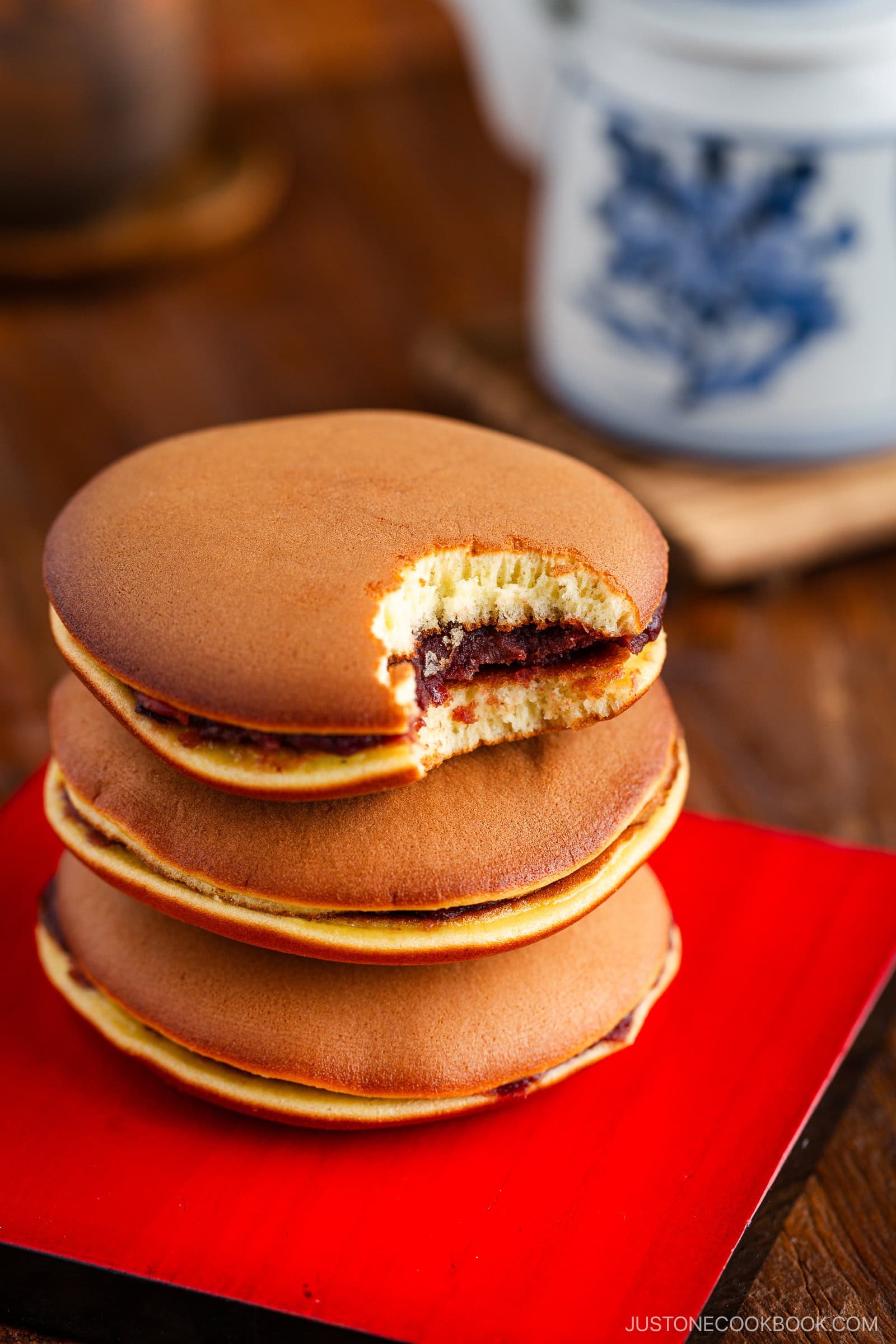 Three dorayaki (Japanese red bean pancakes) are stacked on a red tray, the top one bitten into to reveal its sweet filling. A blue and white teapot is softly blurred in the background.