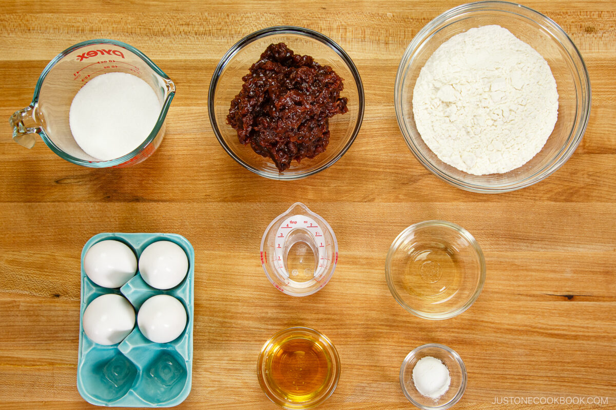 Baking ingredients arranged on a wooden surface for dorayaki (Japanese red bean pancake): sugar, flour, eggs, chocolate mixture, oil, water, vanilla extract, and salt—each neatly placed in separate bowls or measuring cups in an overhead layout.