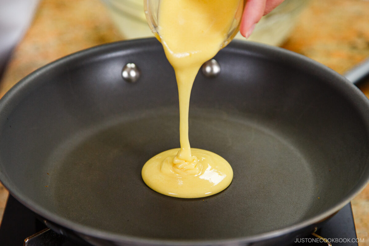 A hand pours thick, yellow dorayaki batter from a glass bowl onto a nonstick frying pan over a stovetop, ready to make the classic Japanese red bean pancake.