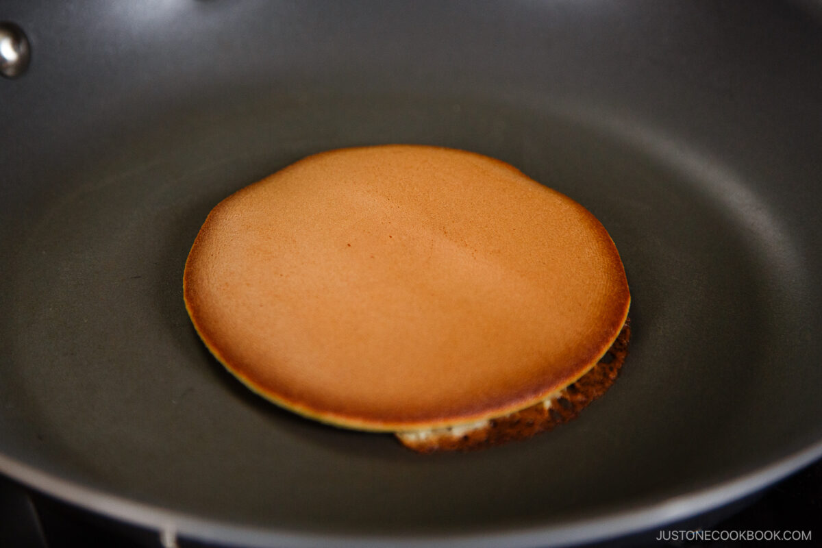A single golden-brown dorayaki, a Japanese red bean pancake, cooking in a nonstick frying pan.