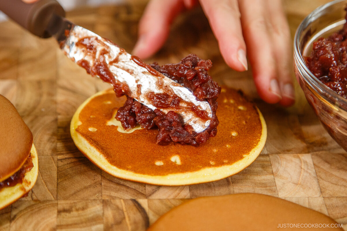 A hand spreads sweet red bean paste onto a round pancake with a knife, preparing dorayaki (Japanese red bean pancake) on a wooden surface. A bowl of red bean paste and extra pancakes sit nearby.