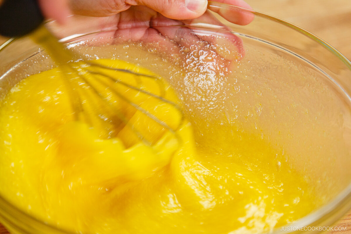 A close-up of a hand whisking bright yellow eggs in a glass bowl, creating a swirling motion as the first step in making dorayaki, the beloved Japanese red bean pancake.