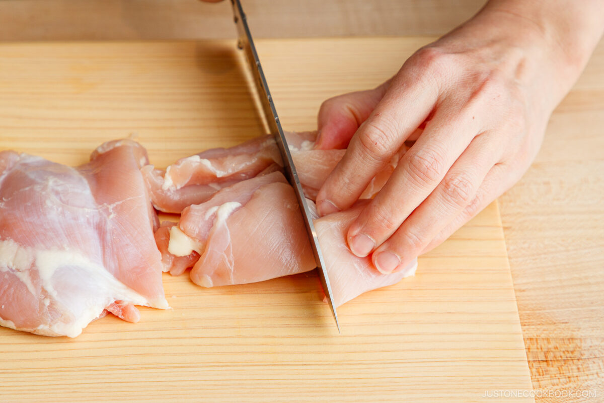 A hand uses a sharp knife to slice raw chicken breast on a light wooden cutting board, preparing ingredients for a Japanese hot pot meal prep.