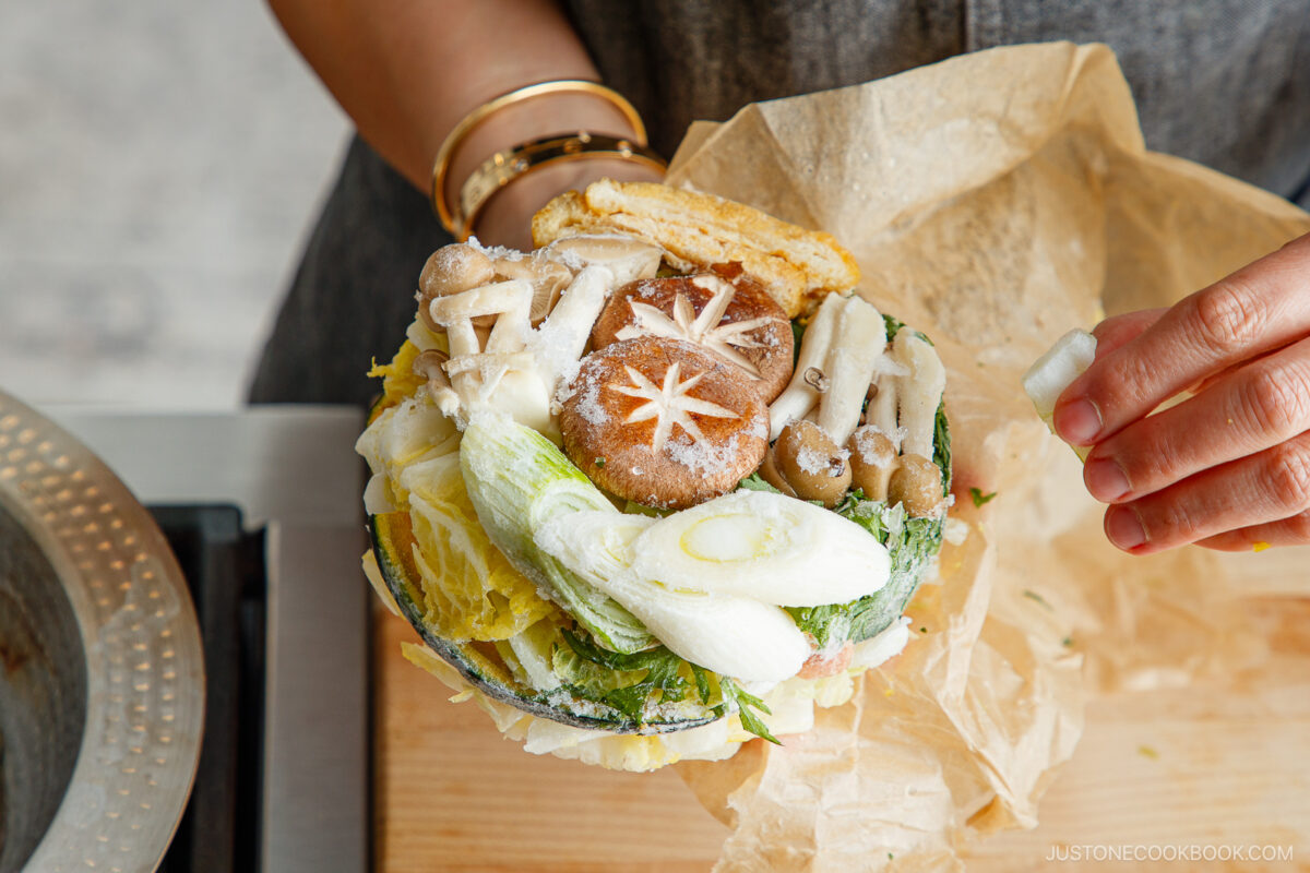 A hand holds a paper-wrapped bundle of assorted fresh vegetables, including mushrooms, napa cabbage, and green onions, arranged in a bowl shape—perfect for Japanese hot pot meal prep and ready for cooking.