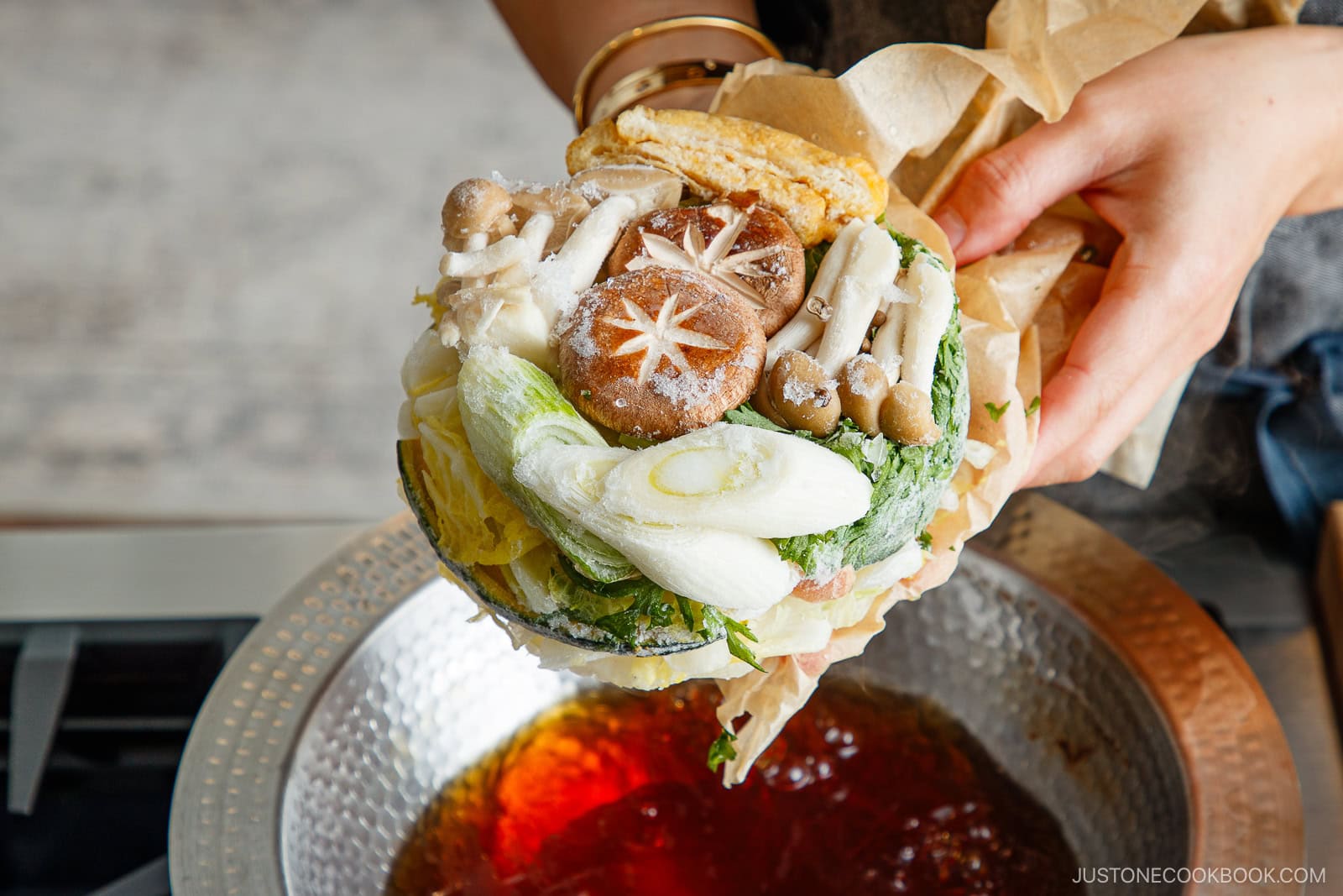 A hand holds parchment paper filled with assorted fresh vegetables and mushrooms, ready to be added to a pot of simmering broth on a stove as part of Japanese hot pot meal prep.