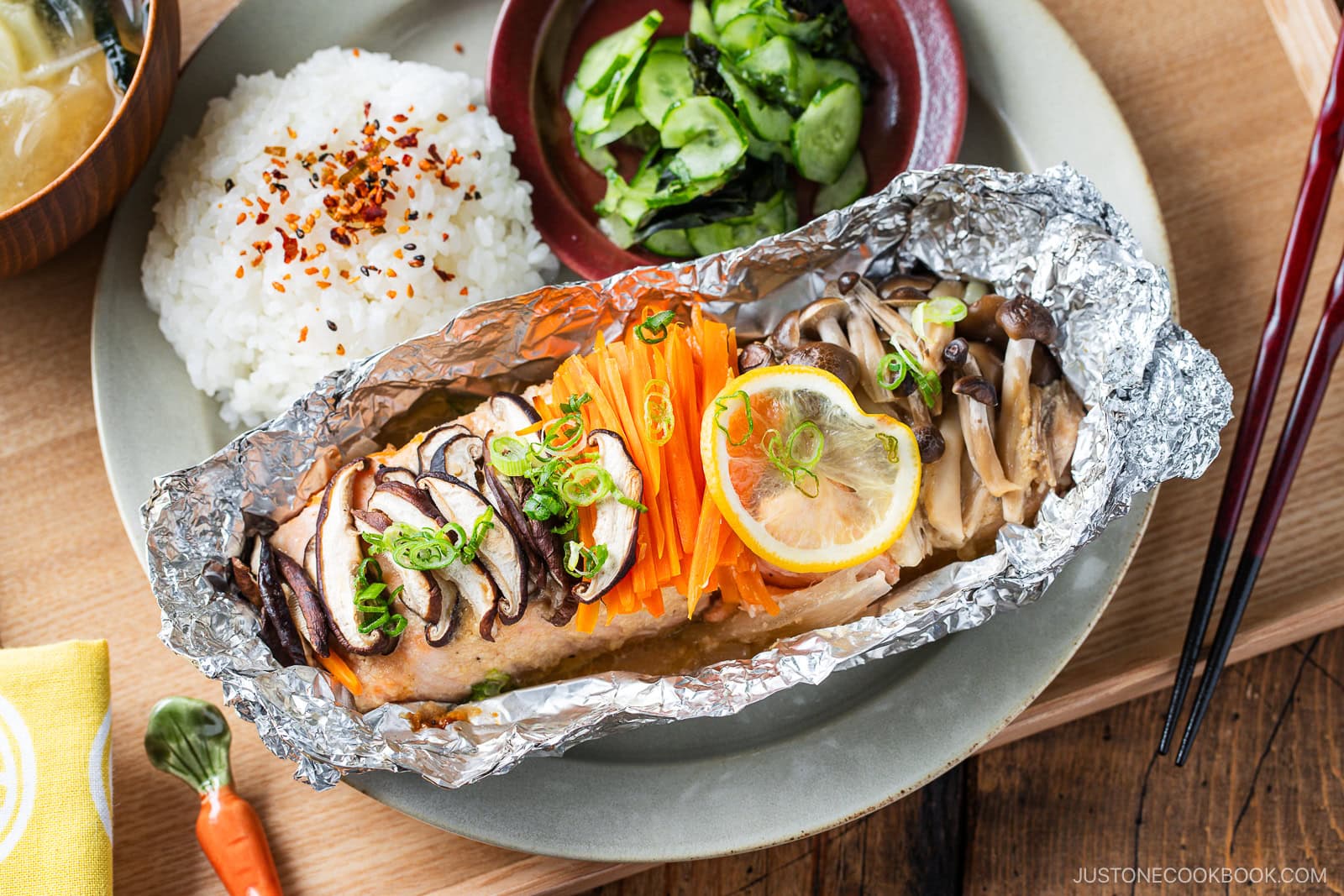 A plate featuring miso salmon in foil, topped with sliced mushrooms, carrots, green onions, and a lemon slice, served alongside steamed white rice and a small bowl of cucumber salad. Chopsticks and a spoon are on the side.