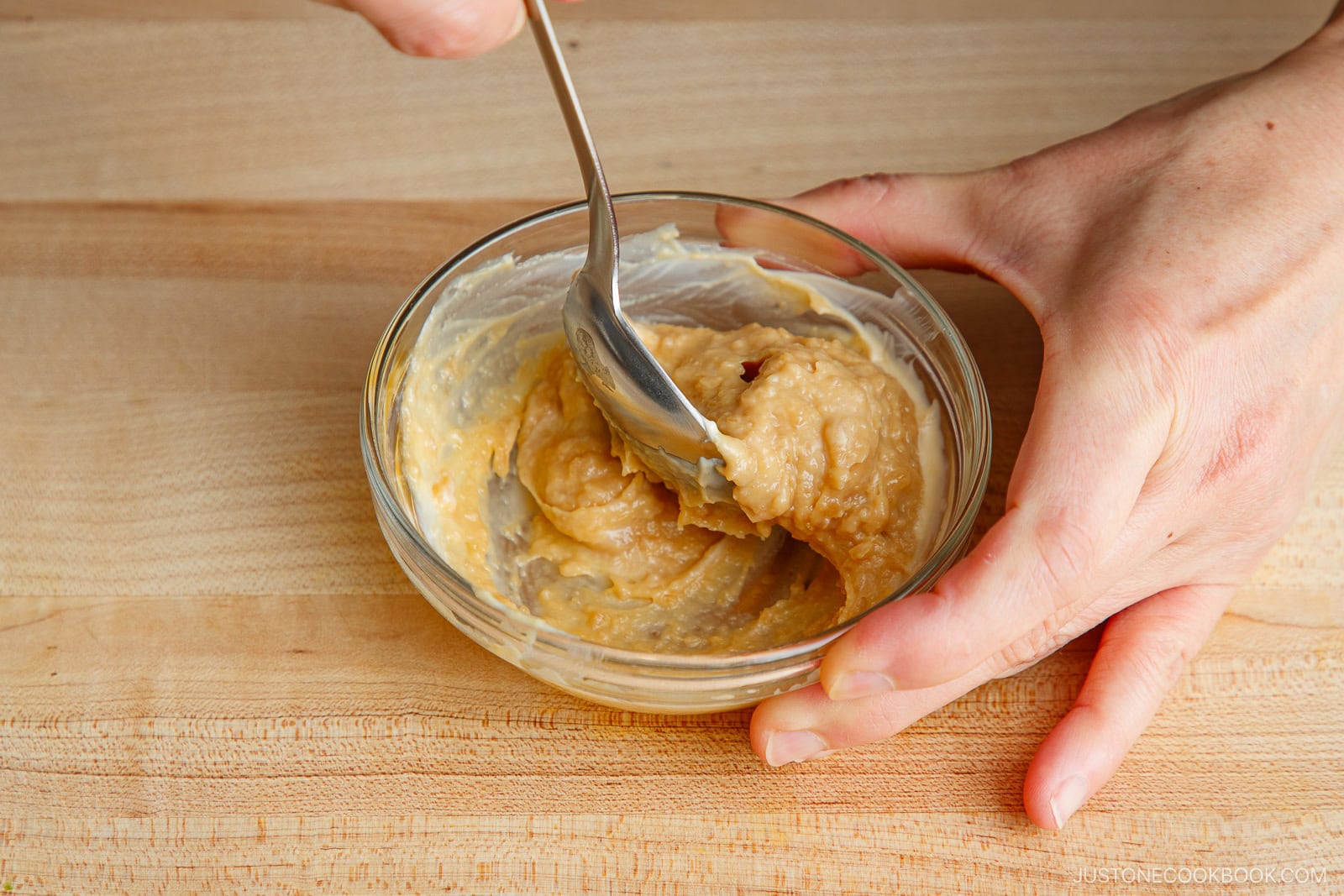 A person mixes a thick, light brown paste in a small glass bowl with a spoon on a wooden surface, preparing the savory marinade for miso salmon in foil.
