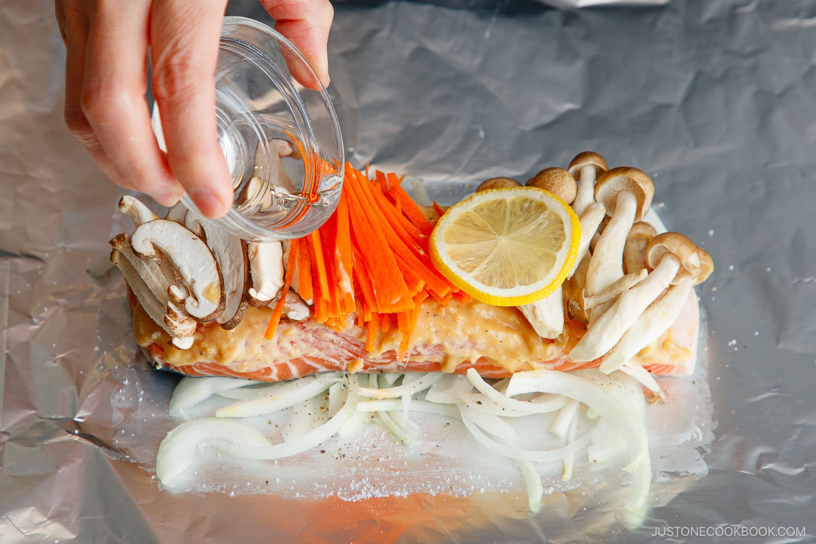 A hand pours liquid from a small glass bowl onto a miso salmon in foil fillet topped with sliced mushrooms, carrots, and lemon on a bed of onions, all arranged on aluminum foil.