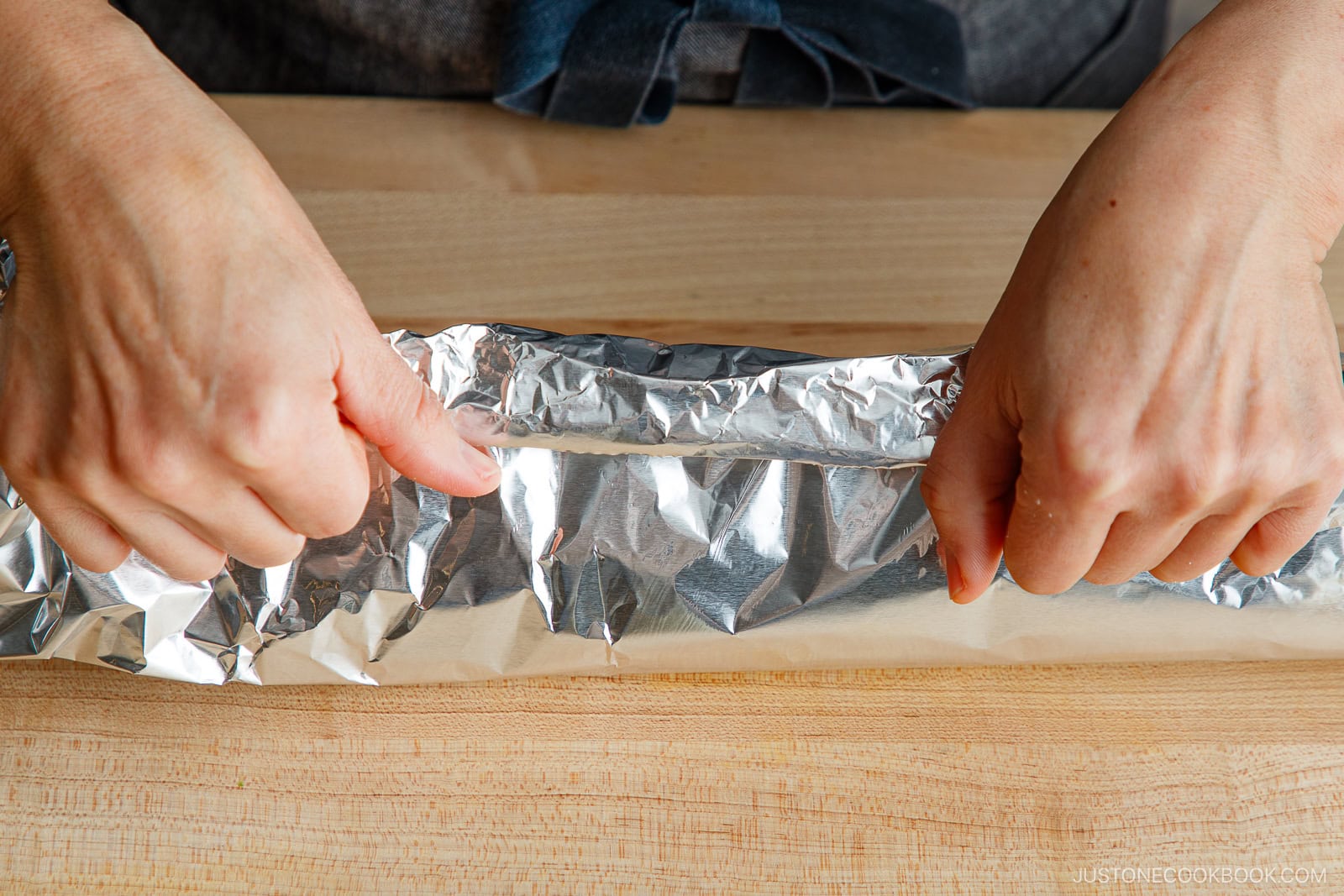 Close-up of hands wrapping a long roll of miso salmon in foil on a wooden surface, sealing in the flavors.