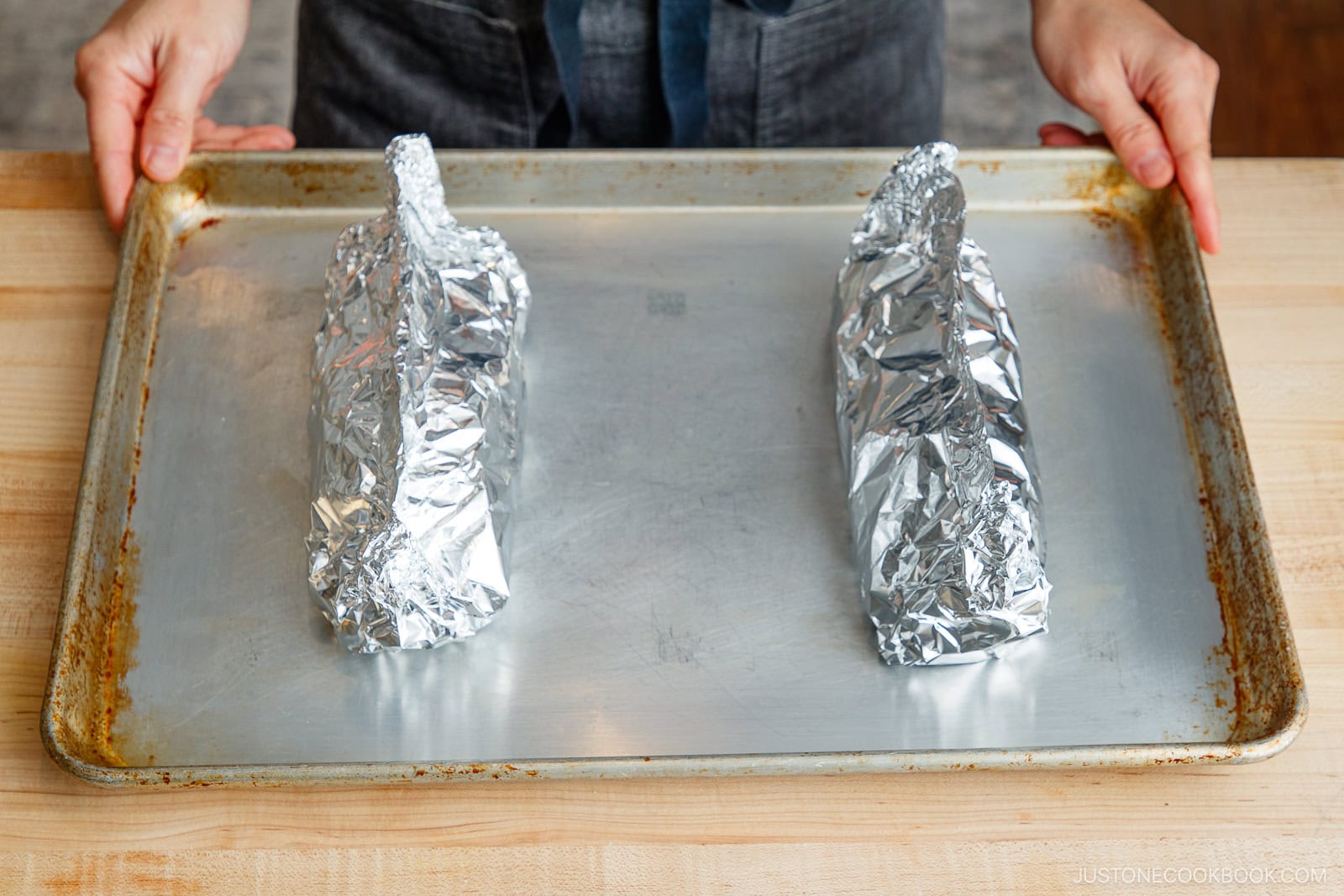 A person places a baking sheet with miso salmon in foil and another food item on a wooden countertop, preparing them for cooking.