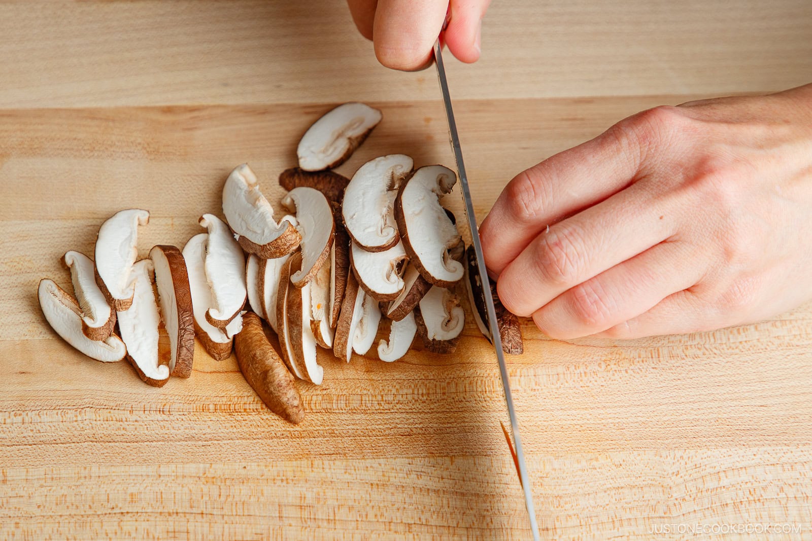 A person’s hand slices shiitake mushrooms on a wooden cutting board with a knife, prepping ingredients for miso salmon in foil.