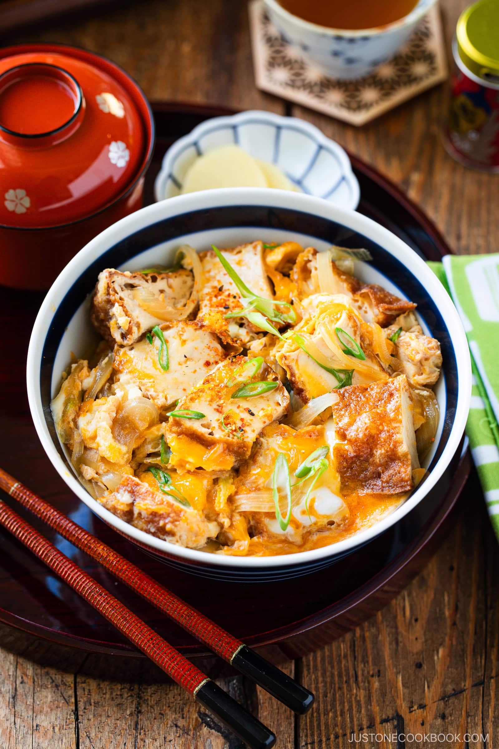 A bowl of Japanese Simmered Tofu and Egg Rice Bowl with fried tofu, egg, onions, and scallions sits on a tray next to red chopsticks, pickled ginger, a lidded bowl, and a cup of tea on a wooden table.