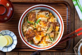 A bowl of Simmered Tofu and Egg Rice Bowl with tofu, softly cooked eggs, and onions simmered in a savory sauce, then spooned over steamed rice—placed on a wooden tray with pickled ginger, chopsticks, and a miso soup bowl nearby.