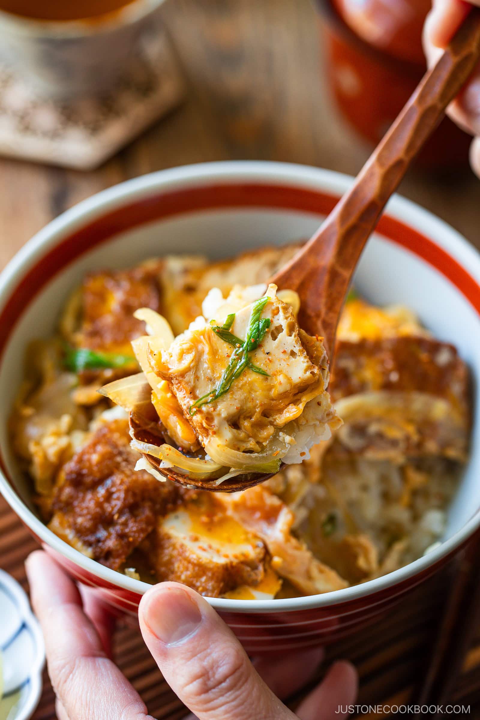 A close-up of a hand holding a wooden spoon with a bite of Simmered Tofu and Egg Rice Bowl—fried tofu, egg, and onions—a reminiscent of comforting classics like oyakodon.