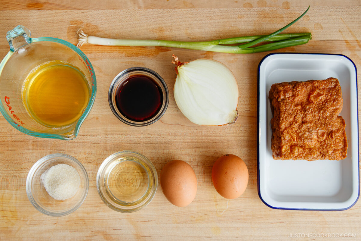 Ingredients on a wooden surface: a measuring cup of broth, soy sauce in a small bowl, a green onion, half a white onion, a plate with fried tofu, sugar, cooking sake in a bowl, and two eggs.