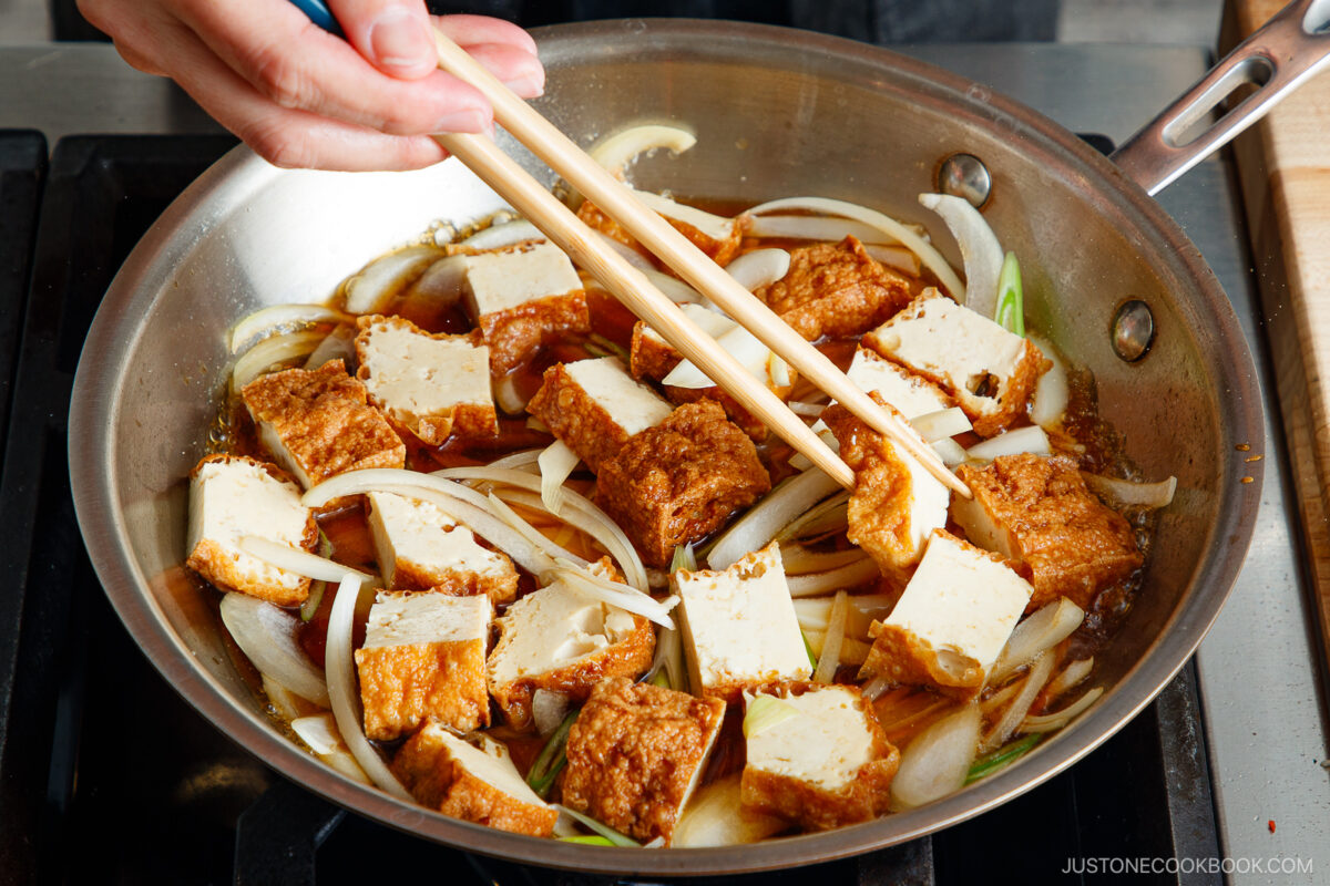 A hand uses chopsticks to cook slices of tofu and onions in a brown sauce on a stovetop, preparing the flavorful base for a Simmered Tofu and Egg Rice Bowl.
