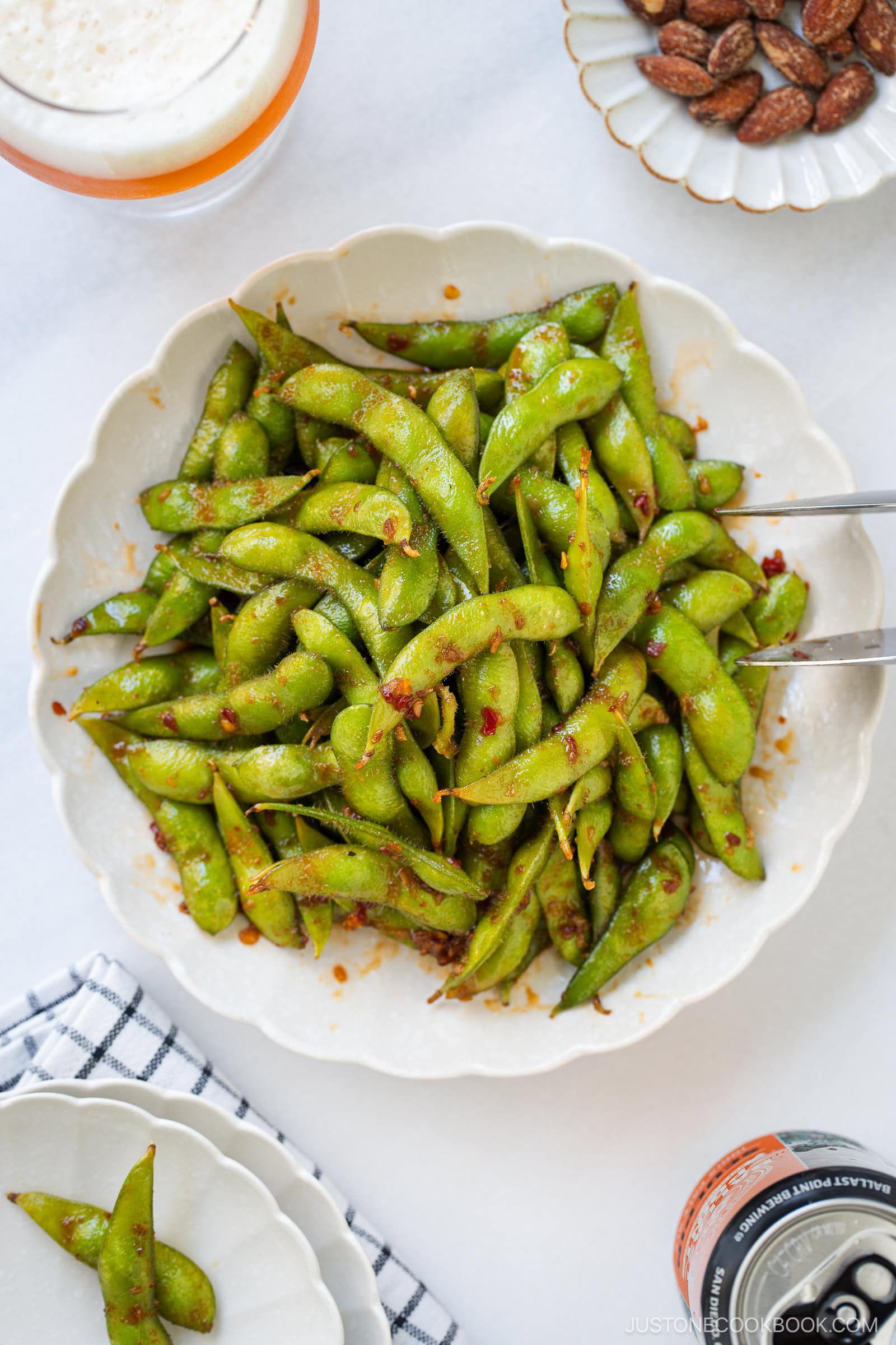 A bowl of spicy edamame with tongs, surrounded by a glass of foam-topped drink, a small bowl of almonds, and a napkin on a white surface.