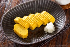Sliced Dashimaki Tamago (Japanese Dashi Rolled Omelette) is arranged neatly on a dark plate with a small portion of grated daikon radish. The plate rests on a wooden table beside a pair of chopsticks.