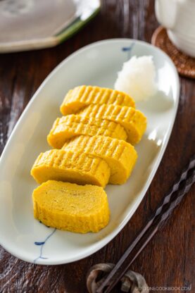 Five slices of Dashimaki Tamago (Japanese Dashi Rolled Omelette) are neatly arranged on a white oval plate, accompanied by a small mound of grated daikon radish. Chopsticks rest beside the plate on a wooden table.
