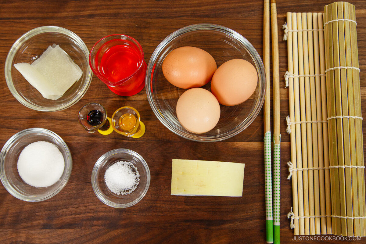 A flat lay of sushi-making ingredients and tools on a wooden surface, featuring essentials for Dashimaki Tamago (Japanese Dashi Rolled Omelette), including eggs, sugar, salt, vinegar, soy sauce, mirin, kombu, a tamagoyaki pan, chopsticks, and a bamboo mat.