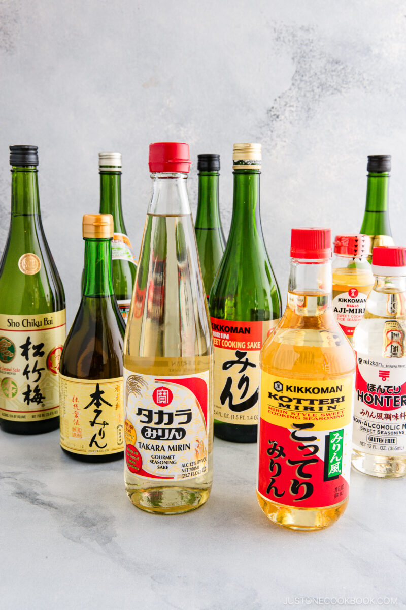 An assortment of Japanese cooking sake, mirin, and rice vinegar bottles arranged on a light gray surface, featuring various brands and colorful labels. Discover Sake vs Mirin: Key Differences and How to Use Them in your kitchen creations.